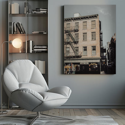 A low-angle, desaturated photograph of a classic brown brick building in a city, featuring multiple windows and a black metal fire escape zigzagging up the side. The sky is a pale, hazy blue, and the ground floor has dark storefronts. Decor