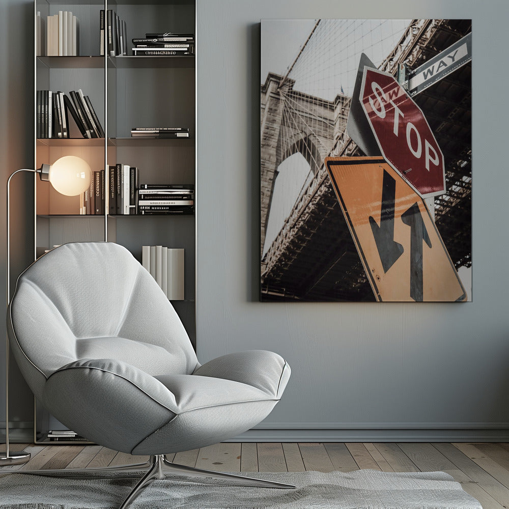 A low-angle photograph of the Brooklyn Bridge with a collection of street signs in the foreground. A red stop sign and a yellow sign with two arrows are prominent against the architectural details of the bridge. Print