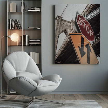 A low-angle photograph of the Brooklyn Bridge with a collection of street signs in the foreground. A red stop sign and a yellow sign with two arrows are prominent against the architectural details of the bridge. Print