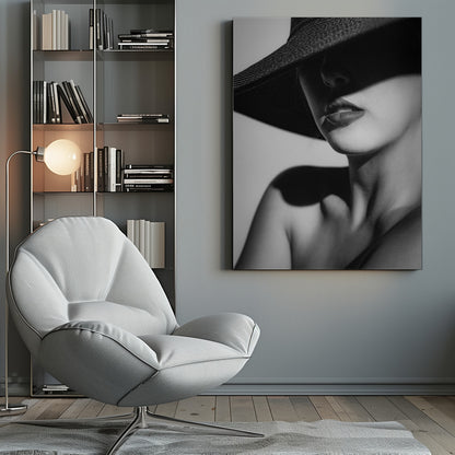A dramatic black and white close-up photograph of a woman wearing a large, dark, wide-brimmed hat. The hat casts a deep shadow over the upper half of her face, obscuring her eyes and creating a sense of mystery. Only her nose, full lips, chin, neck, and shoulder are visible, highlighted by a strong light source. Decor