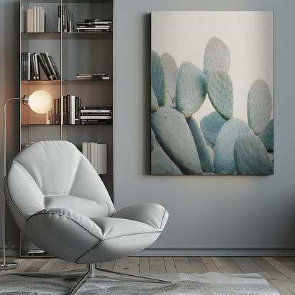 A close-up photograph of pale, dusty green prickly pear cactus pads against a bright, off-white background, presented in a minimalist style with a thin black frame. Artwork