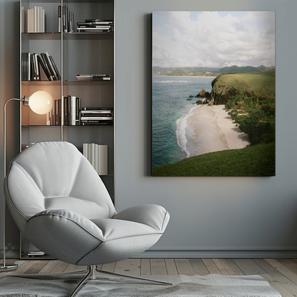 A high-angle photograph of a secluded tropical beach, with turquoise waves gently washing onto the white sand. A lush green cliff covered in grass and small trees frames the cove, with distant rolling hills visible under a soft, cloudy sky. Decor