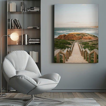 A wooden staircase and walkway descends a green, scrub-covered hill towards a rocky point overlooking a churning blue ocean under a pale, hazy sky. Print