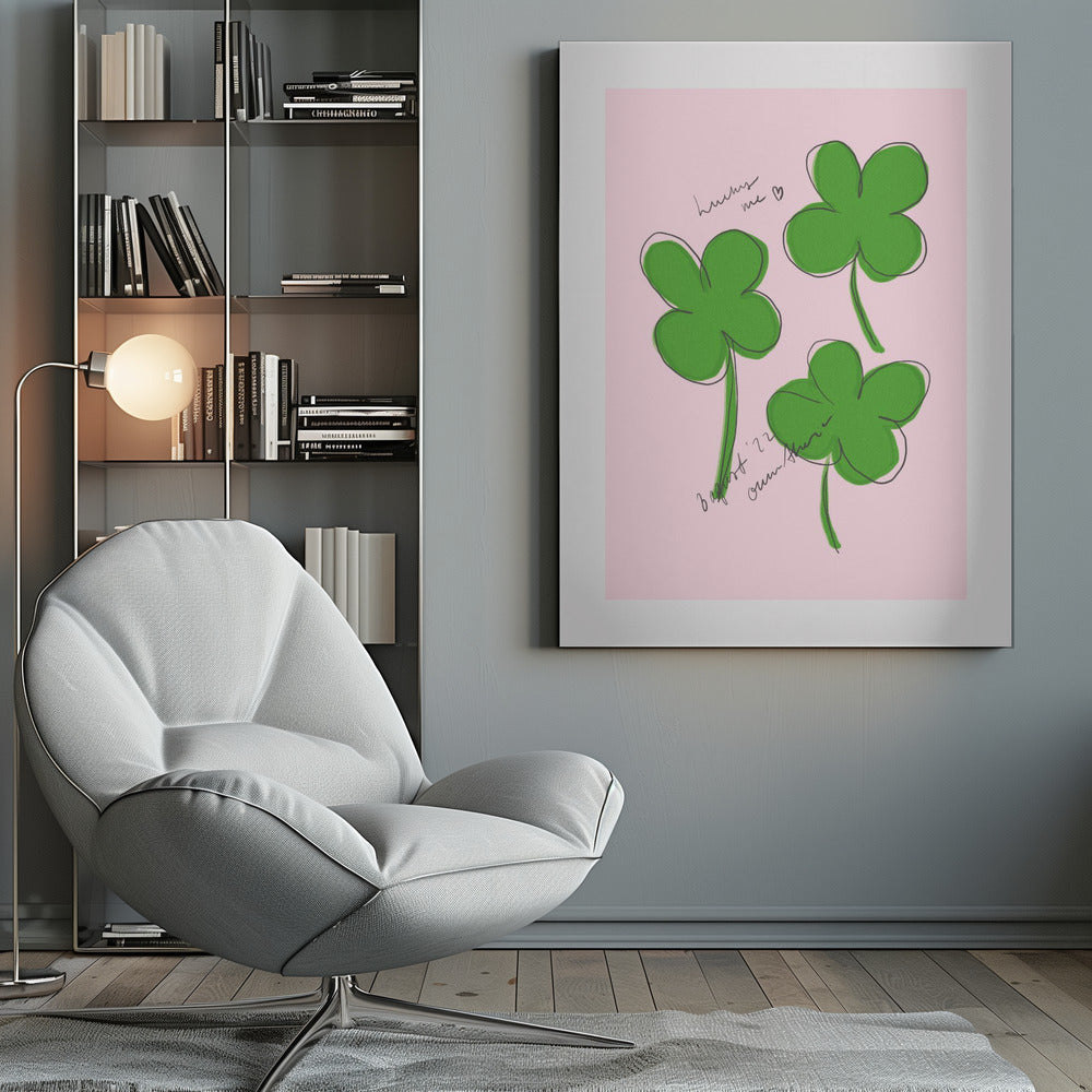 A framed art print featuring three hand-drawn four-leaf clovers in green with a loose black outline, set against a solid light pink background. Cursive handwriting on the print reads 'lucky me ♡' and includes a signature. Artwork