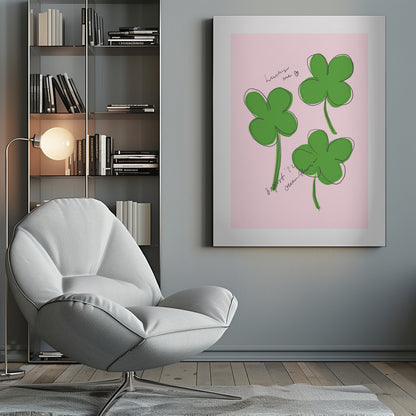 A framed art print featuring three hand-drawn four-leaf clovers in green with a loose black outline, set against a solid light pink background. Cursive handwriting on the print reads 'lucky me ♡' and includes a signature. Artwork