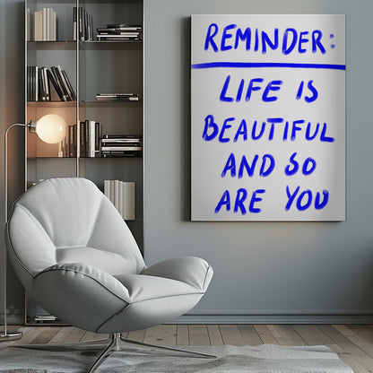 A framed poster featuring a positive affirmation handwritten in a blue, brush-stroke style font on a white background. The text reads 'REMINDER: LIFE IS BEAUTIFUL AND SO ARE YOU', with a blue line separating 'REMINDER:' from the rest of the quote. Print