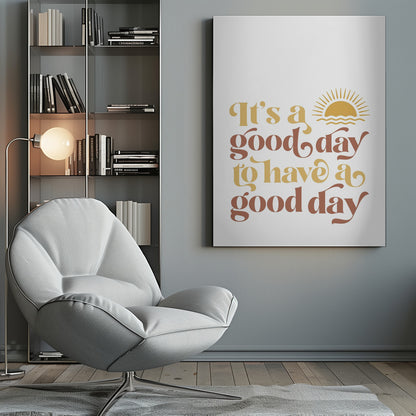 A framed art print with a white background featuring the quote 'It's a good day to have a good day' in a retro, curvy font. The text color alternates between terracotta brown and mustard yellow. Above the text is a small yellow icon of a sun rising over waves. Wall Art