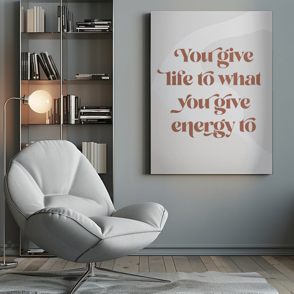 A framed motivational poster featuring the quote 'You give life to what you give energy to'. The text is printed in a dusty rose color using a stylish, retro serif font on a light gray background with subtle wavy shapes. Poster