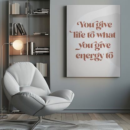 A framed motivational poster featuring the quote 'You give life to what you give energy to'. The text is printed in a dusty rose color using a stylish, retro serif font on a light gray background with subtle wavy shapes. Poster