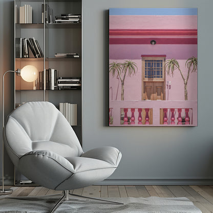 A brightly colored pink building stands under a clear blue sky, framed in black. A central wooden door with a paned window is flanked by two slender, palm-like trees. In the foreground, a pink balcony wall features fuchsia-colored balusters. Decor