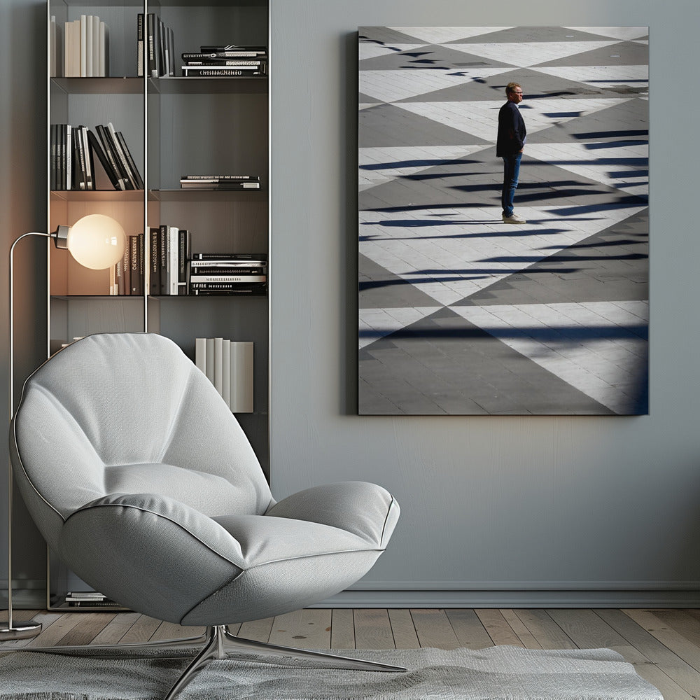 A man in a dark jacket and blue jeans stands alone in profile on a geometrically patterned plaza. The ground is a striking pattern of grey and white triangles, crossed by long, dark shadows cast by the bright sun, creating a high-contrast, graphic composition. Wall Art