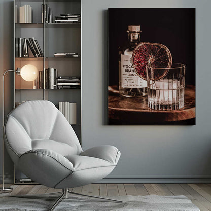 A moody still life photograph of a cocktail in a cut-crystal glass filled with ice. A dried blood orange slice garnishes the rim. In the soft-focus background, a bottle of gin rests on a dark wooden tray against a black backdrop. Wall Art