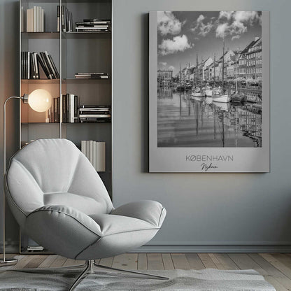 A black and white poster of Nyhavn harbor in Copenhagen. The photograph shows traditional sailboats and ships moored along a canal, with a line of historic, multi-story buildings on the far bank. The sky has fluffy clouds, and the entire scene is reflected in the calm water. The poster has a white border with the text 'KØBENHAVN', 'Nyhavn', and 'DANMARK' at the bottom, and is shown within a black frame. Poster