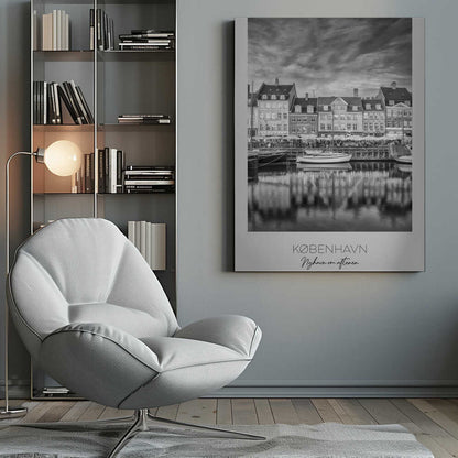 A framed black and white photograph of the Nyhavn harbor in Copenhagen, Denmark. The image shows a row of classic gabled houses along the waterfront, with several sailboats moored in the canal. The buildings and a dramatic cloudy sky are reflected in the water. At the bottom, a white border contains the text 'KØBENHAVN', 'Nyhavn om aftenen', and 'DANMARK'. Wall Art