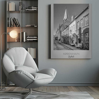 A black and white framed travel poster of a charming cobblestone street in Nyboder, Copenhagen. The photograph shows a sunlit row of historic houses with a prominent church steeple in the background. At the bottom of the poster, the words 'KØBENHAVN', 'Nyboder', and 'DANMARK' are written. Wall Art