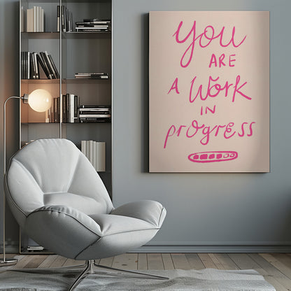 A framed art print featuring the motivational quote 'You are a work in progress'. The text is rendered in a vibrant pink, hand-lettered style against a soft beige background. The words 'You', 'a Work', and 'progress' are in a flowing script, while 'are' and 'in' are in a simple uppercase font. Wall Art