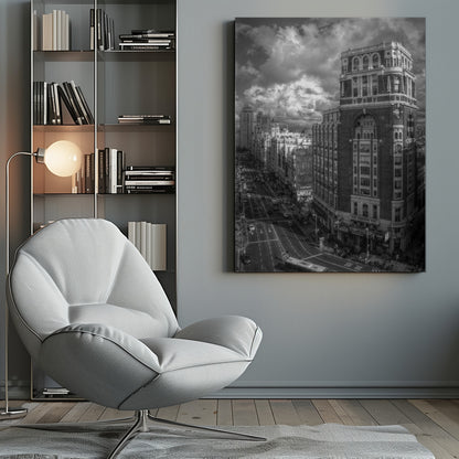 A dramatic black and white high-angle photograph of a bustling city street. A large, ornate historical building dominates the right side, while traffic flows along the avenue on the left under a sky filled with heavy, textured clouds. Poster