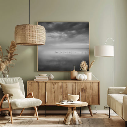 A minimalist black and white photograph in a light wood frame, depicting a calm, serene lake under a heavy, cloudy sky. The still water reflects the sky, with a row of weathered wooden posts breaking the surface along the horizon. Print