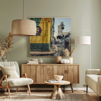 A framed photograph of a vibrant street corner in a city like Havana, Cuba, with a large mural of Che Guevara on a yellow and green wall. The wet street reflects the buildings and a palm tree, while people, a vintage car, and a rickshaw occupy the narrow road. Poster