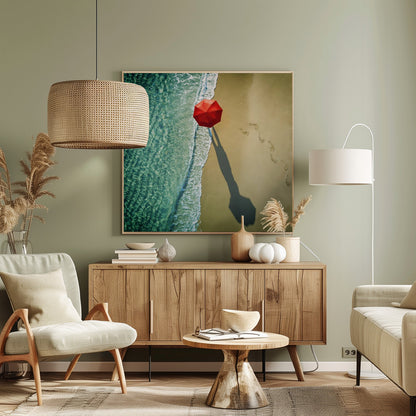 An aerial photograph of a serene beach scene, featuring a vibrant red umbrella on the golden sand next to the clear turquoise ocean. Gentle white waves lap the shore, and a long shadow from the umbrella stretches across the sand, where a trail of footprints leads away. Poster