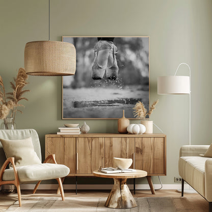 A close-up, black and white photograph of a ballerina's feet in worn pointe shoes, captured in mid-air above a puddle, with water splashing upwards. The motion is frozen, highlighting the contrast between grace and a raw outdoor element. The image is presented in a light wood frame. Poster