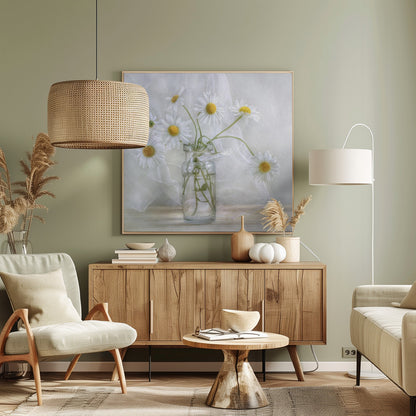 A delicate still life photograph of several white daisies with bright yellow centers in a clear glass jar. The arrangement sits on a light wooden surface against a backdrop of soft, draped white fabric. The entire image is enclosed in a light wood frame. Artwork