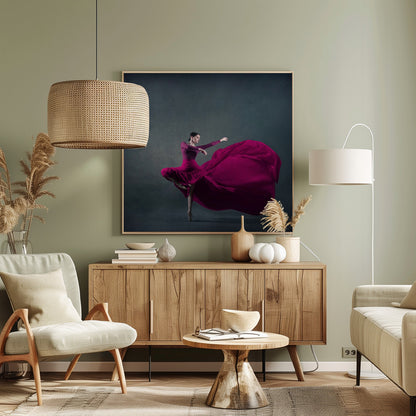 A graceful ballet dancer in a vibrant magenta dress strikes a pose on pointe, with the long, flowing fabric of her skirt billowing out dramatically behind her against a dark, textured gray background, all enclosed in a simple light wood frame. Decor