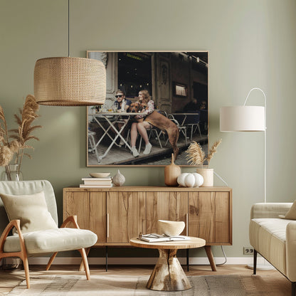 A framed photograph of a young couple sitting at an outdoor cafe. A woman in a floral shirt holds a large brown Rhodesian Ridgeback on her lap, while a man in sunglasses sits beside her. The dog stands with its front paws on the woman's lap and back paws on the wooden deck. The scene has a muted, warm tone. Poster