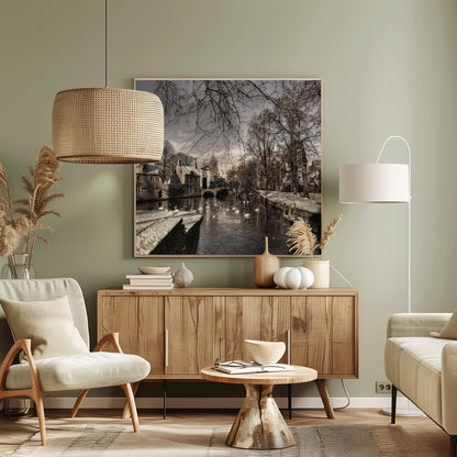 A vintage-style, framed photograph of a European canal scene in winter. Historic buildings and bare trees line the partially frozen canal, where several white swans are swimming. A light dusting of snow covers the canal banks and rooftops under a cloudy sky. Wall Art