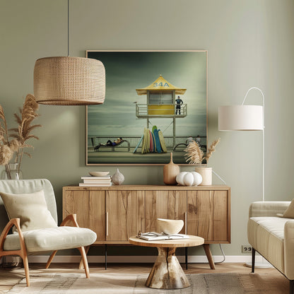 A vintage-style photograph of a yellow lifeguard tower labeled '34A' on a beach boardwalk under an overcast, greenish sky. A lifeguard in a wetsuit stands on the balcony, while a collection of colorful surfboards leans against the base. People are seen relaxing on benches nearby. Print