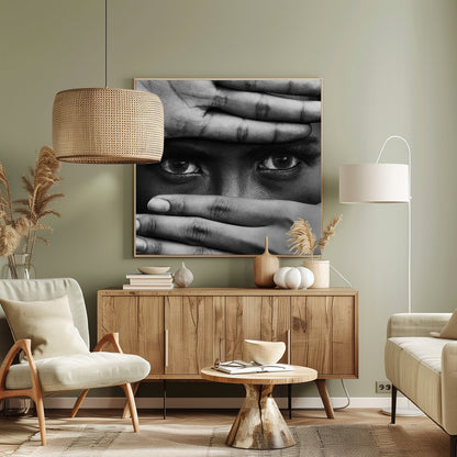 A powerful black and white close-up photograph of a person's face, framed in light wood. Their hands cover most of their face, one over the forehead and the other over the mouth, revealing only their intense, striking eyes that gaze directly forward. Print