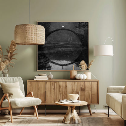 A framed black and white photograph of a ballerina in a white tutu standing at the peak of a semi-circular stone bridge. The bridge and the ballerina are perfectly reflected in the still water below, creating a full circle. The scene is set in a dark, misty forest, giving it a surreal and moody atmosphere. Artwork