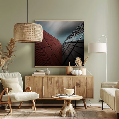 A dramatic low-angle photograph of two modern buildings with contrasting facades. On the left, a building is covered in deep red square panels. On the right, a darker building features black angular tiles and glass windows. The sky above is a moody, blurred grey. Print