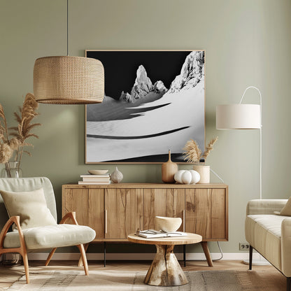 A framed, high-contrast black and white photograph of a majestic, snow-covered mountain range. Jagged peaks are sunlit against a stark black sky, casting long, dramatic shadows across a vast, pristine snowfield below. A tiny, solitary figure stands in the snow, highlighting the immense scale of the landscape. Print