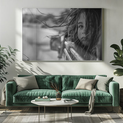 A close-up, black and white photograph of a young girl looking out of a moving vehicle. Her long hair is blowing wildly in the wind, partially obscuring her face as she peers intently at the camera with one eye. Her hand grips the edge of the open window. The background is blurred with motion, suggesting a journey. The photo is displayed in a silver frame. Poster