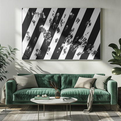 A striking black and white aerial photograph of dozens of people walking across a large, diagonally striped crosswalk. The high-contrast, graphic pattern of black and white stripes dominates the scene, with the small figures of pedestrians scattered across it, creating a sense of movement and urban anonymity. The image is presented within a simple silver frame. Print