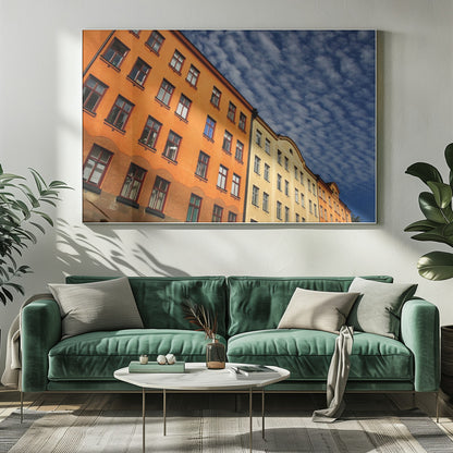 A low-angle shot of colorful buildings, predominantly a vibrant orange one, against a bright blue sky filled with a pattern of white, fluffy clouds. The image is presented within a silver-colored frame. Print