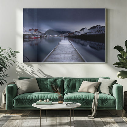 A long exposure photograph of a serene coastal town at night, viewed from a wooden pier that leads into calm, dark water. A lone person stands at the end of the pier, gazing at the town's reflected lights. Behind the village, a mountain looms under a breathtaking, star-filled sky. The image is presented within a silver-colored frame. Artwork