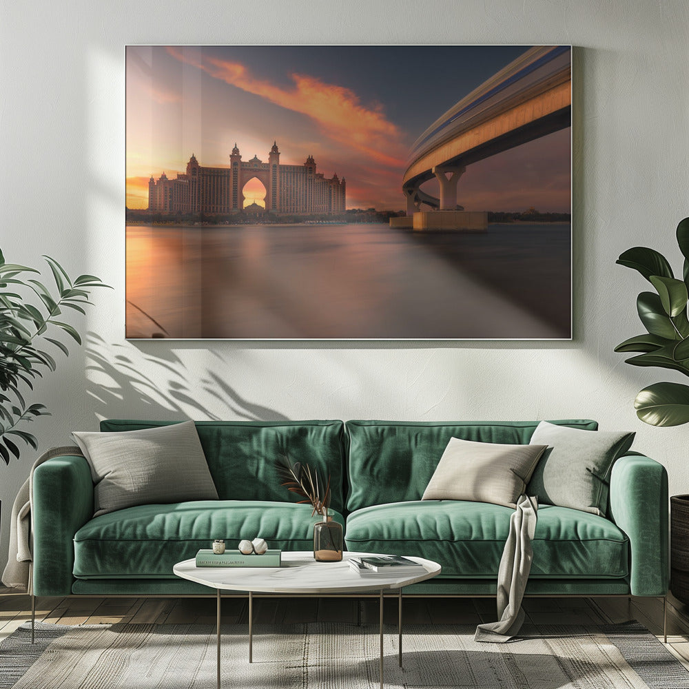 A scenic view of the Atlantis The Palm hotel in Dubai at sunset, with its grand architecture silhouetted against a vibrant orange and pink sky. A modern monorail track curves over the water on the right, with a blurred train indicating motion. The calm water in the foreground reflects the warm colors of the dusk sky. Decor