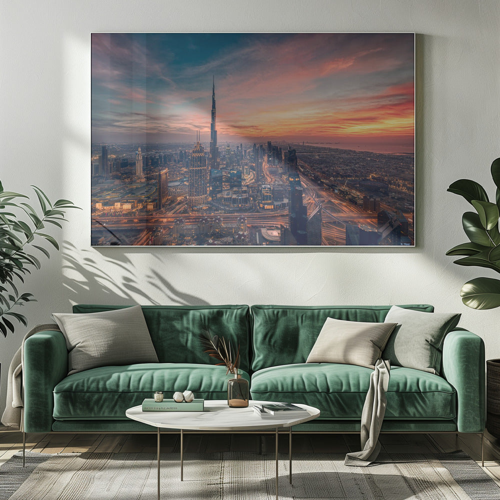 An aerial view of the Dubai skyline at sunset, with the Burj Khalifa prominently featured against a dramatic sky filled with orange, pink, and blue clouds. The sprawling city below is illuminated with golden lights from buildings and busy highways. Decor