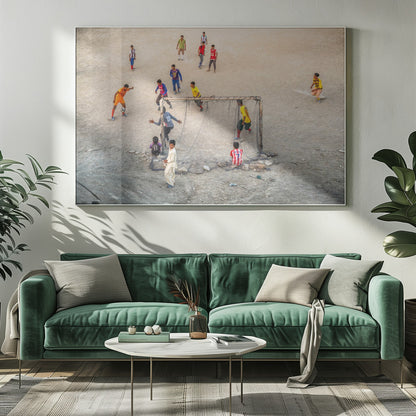 An overhead view of a group of boys and young men playing a spirited game of soccer on a large, dusty dirt field. A player in a bright orange uniform prepares to kick the ball towards a makeshift goal, defended by a goalie in a dark blue jersey. Other players in various colored shirts are scattered across the field, while a few young spectators watch from the sidelines. The entire image is enclosed in a silver frame. Wall Art