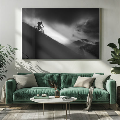 A striking black and white photo of a mountain biker's silhouette on a steep decline, set against a dramatic, light-filled cloudy sky with distant, misty mountains. Artwork