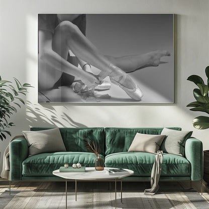 A black and white close-up photograph of a ballet dancer sitting on the floor, carefully tying the ribbons of a satin pointe shoe on her right foot. The image has a simple silver frame and captures a quiet moment of preparation. Poster