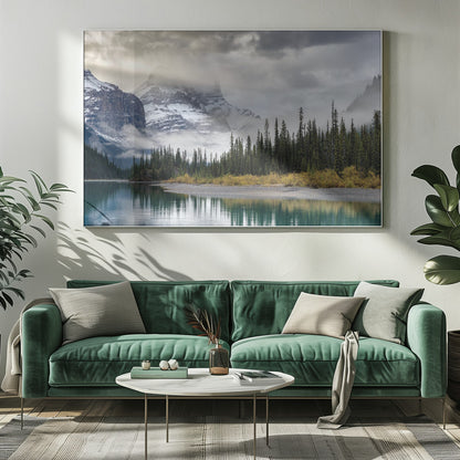 A tranquil mountain landscape featuring a still, teal lake reflecting a dense evergreen forest along its shore. In the background, majestic snow-covered peaks are shrouded in mist and dramatic gray clouds. Wall Art