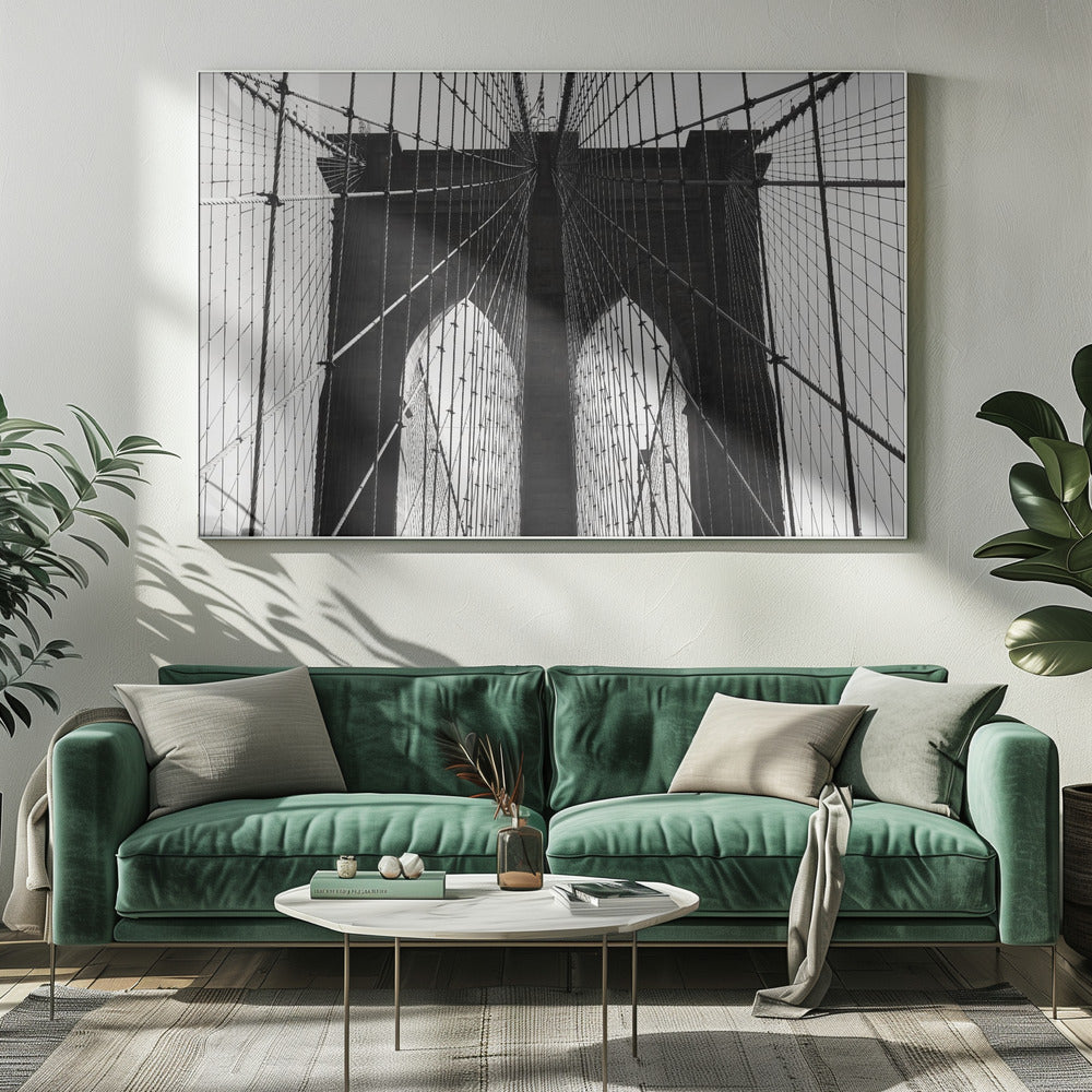 A low-angle, black and white photograph looking up at the iconic stone towers of the Brooklyn Bridge. The intricate web of suspension cables and vertical suspender ropes creates a symmetrical, geometric pattern against the light sky. The photo is enclosed in a silver frame. Print