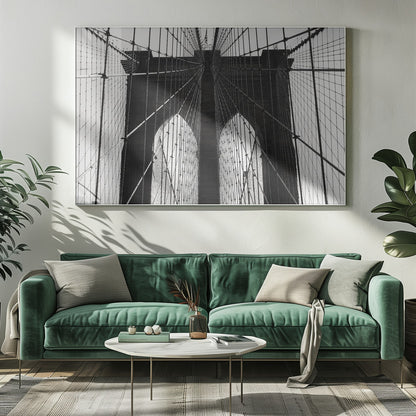 A low-angle, black and white photograph looking up at the iconic stone towers of the Brooklyn Bridge. The intricate web of suspension cables and vertical suspender ropes creates a symmetrical, geometric pattern against the light sky. The photo is enclosed in a silver frame. Print