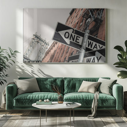 A low-angle shot of a black and white 'ONE WAY' street sign in the foreground, with a red brick building and the Manhattan Bridge visible in the background under an overcast sky, all enclosed in a silver frame. Artwork
