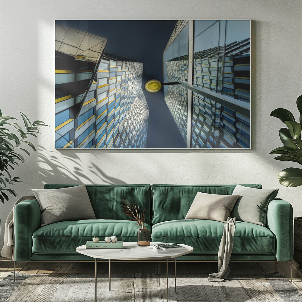 A worm's-eye view looking up between two modern glass skyscrapers with geometric blue, yellow, and silver panels, creating a symmetrical reflection against a dark blue sky, with a bright yellow streetlamp centered in the gap. Decor