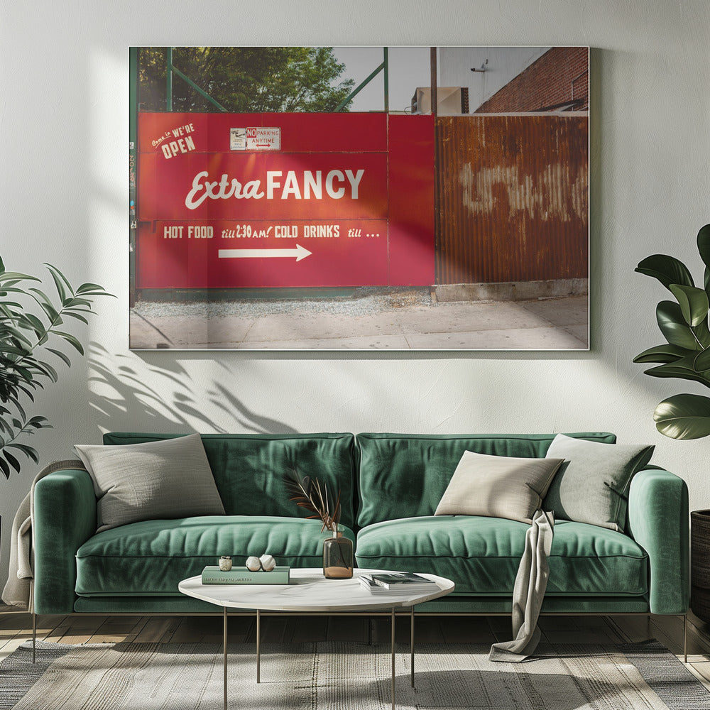 A framed photograph of a bright red outdoor sign for a restaurant named 'Extra Fancy'. The sign, painted with white retro-style text, advertises hot food and cold drinks and is situated next to a rusty, corrugated metal fence on a city sidewalk. Artwork