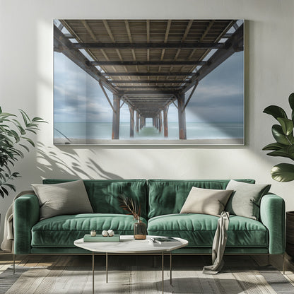 A symmetrical, low-angle photograph taken from underneath a long pier, looking out to sea. The rusty metal pilings and wooden planks of the pier create a receding perspective over the smooth, long-exposure water, all under a cloudy sky and presented within a silver frame. Poster